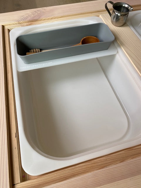 An accessory board for sensory bins holds wooden spoons