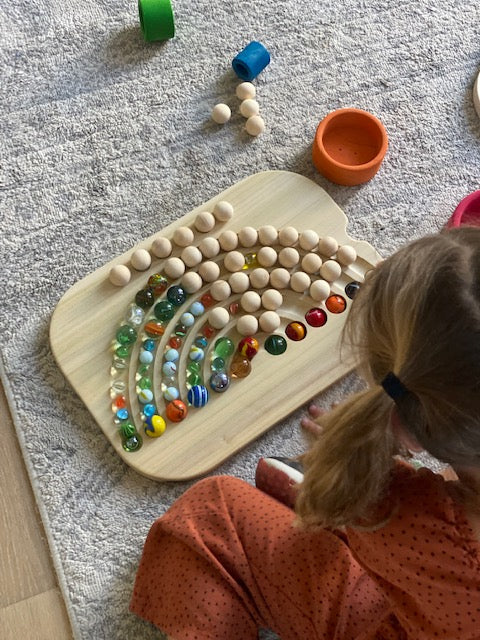 A wooden rainbow board with marbles sits on the ground as a child plays